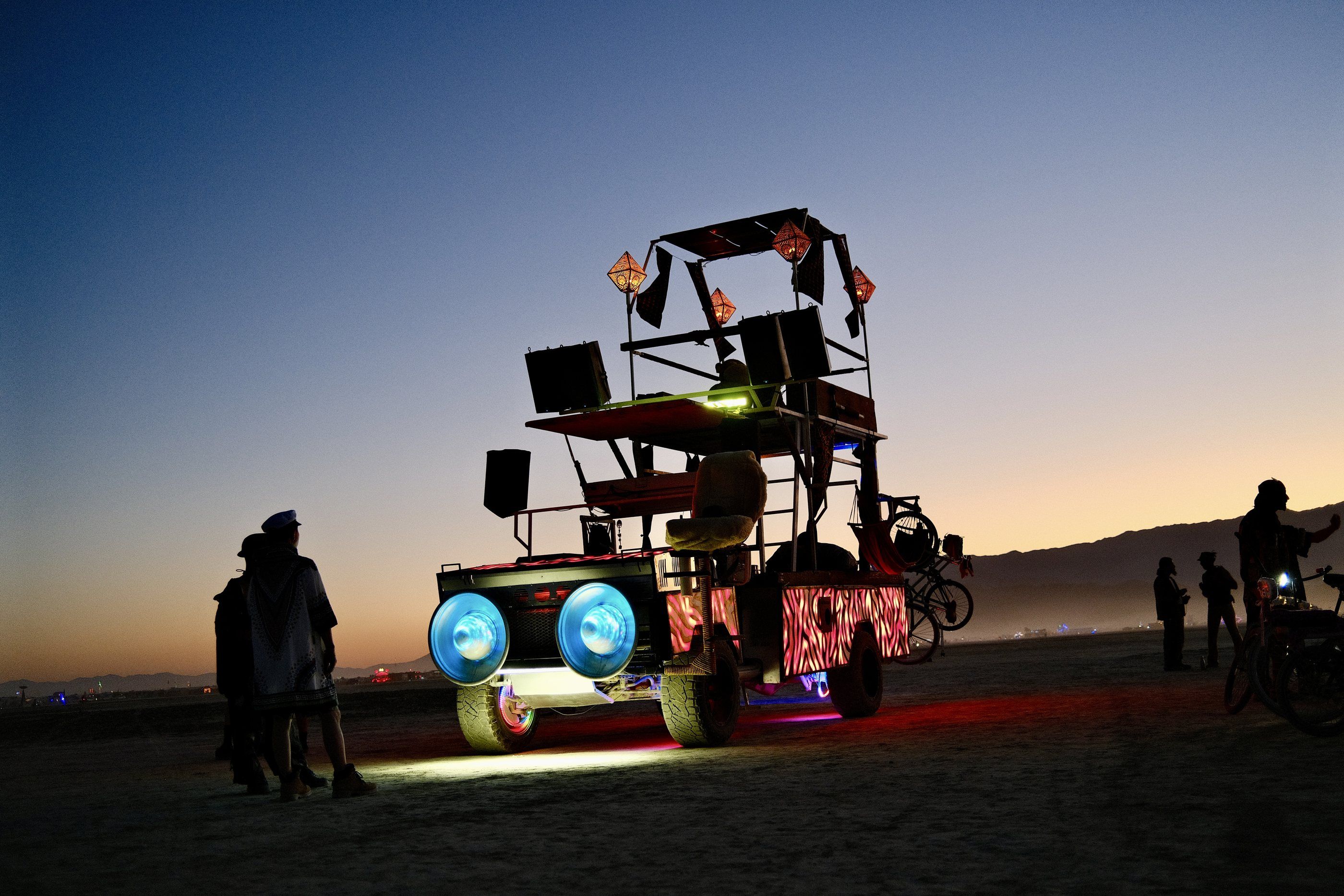 Rexan at dusk on the playa, headlight eyes glowing blue, speakers and lanterns on the top deck